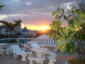 Una terraza con sillones y mesas blancas, con vistas a una puesta de sol sobre una masa de agua y edificios al fondo.