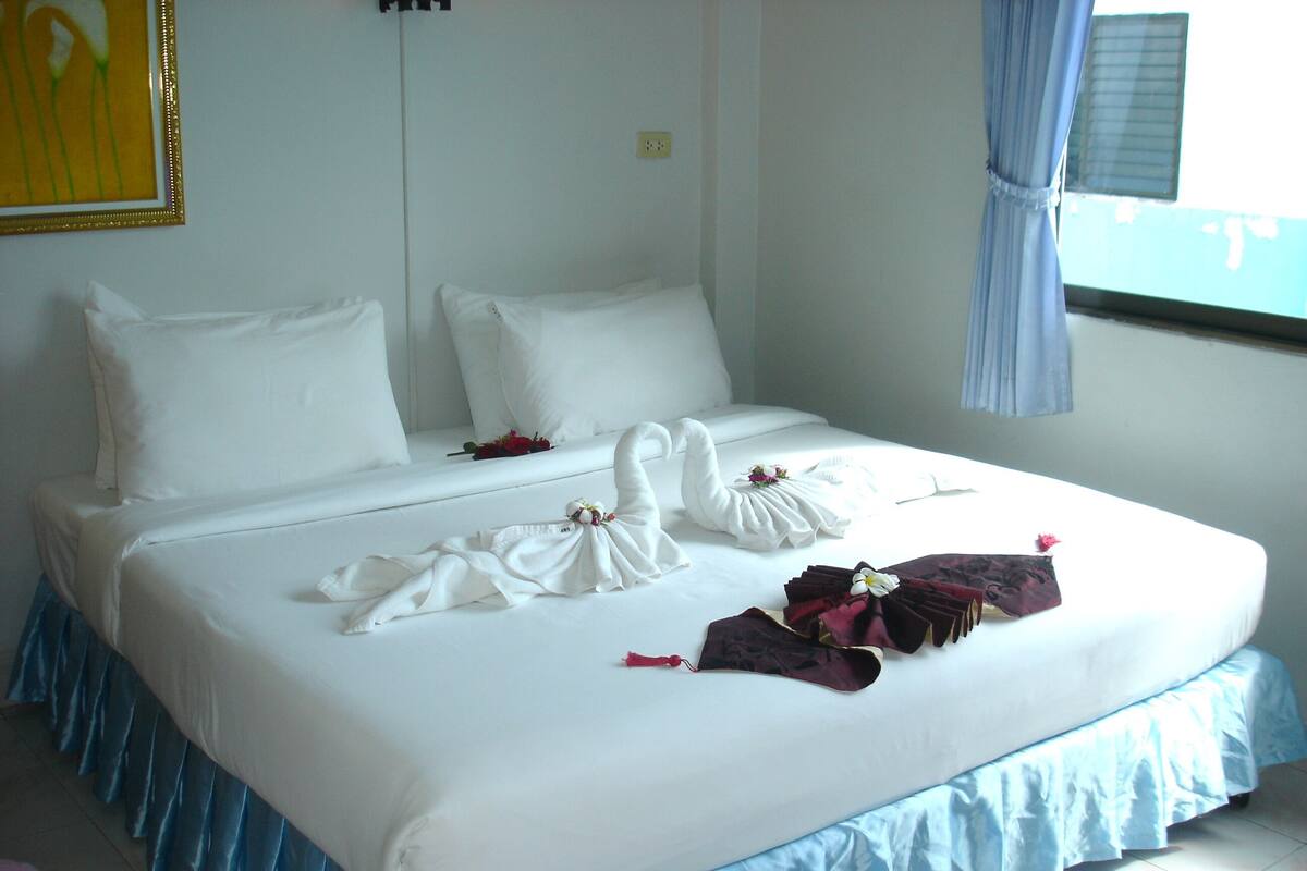 A neatly made bed with white linens and decorative swan-shaped towels, a blue skirted bed base, and a window with curtains.