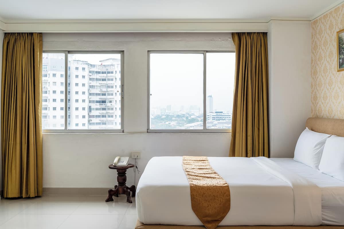 Habitación de hotel con una cama grande, dos ventanas con cortinas, un teléfono en una mesita auxiliar y vistas a un paisaje urbano.