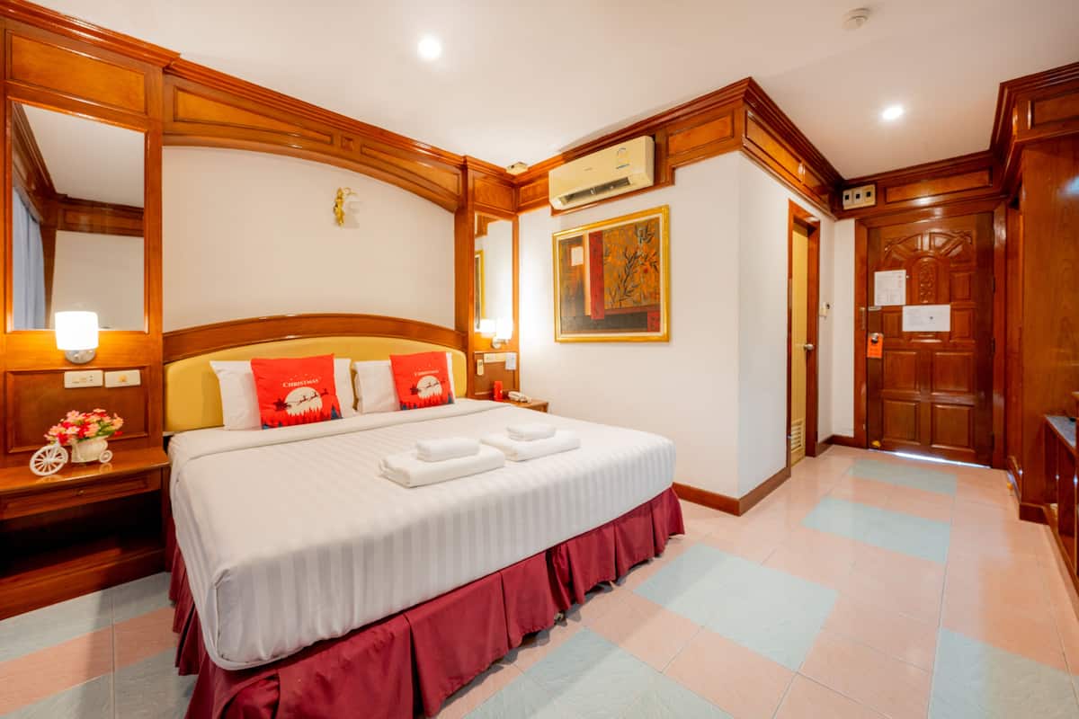 A hotel room with a large bed, wooden headboard, and a red skirt. There is a nightstand with a lamp and a flower vase. The room has a wooden door and a framed picture on the wall.
