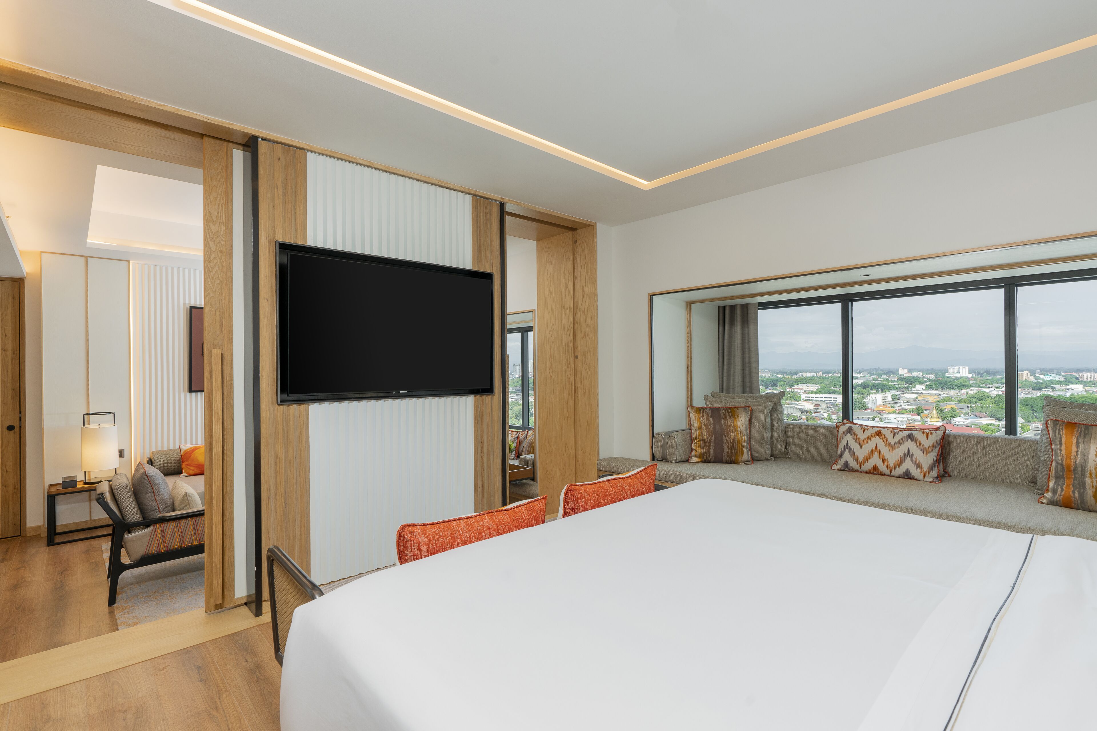 Una habitación de hotel moderna con una cama grande, un televisor montado en la pared, un área de descanso con cojines y vistas a la ciudad a través de amplias ventanas.