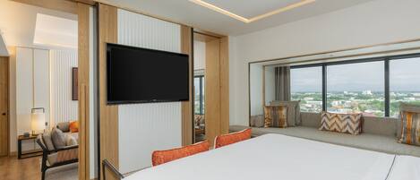 A modern hotel room with a large bed, a TV mounted on the wall, a seating area with cushions, and a view of the city through large windows.