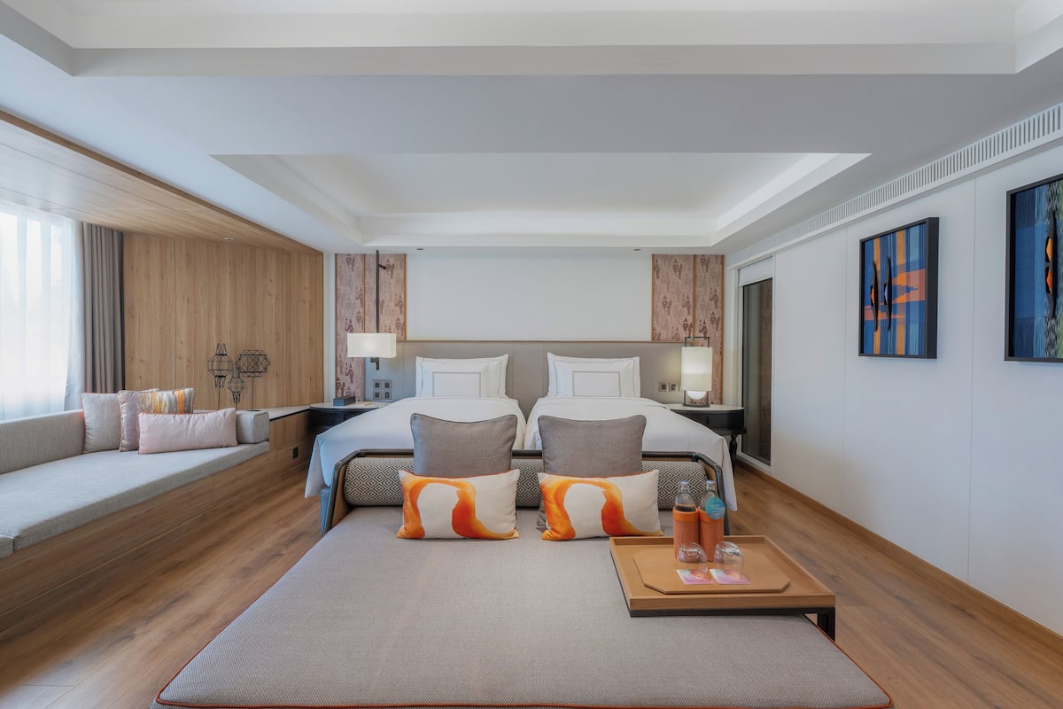 Ein modernes Hotelzimmer mit einem großen Bett, einer Sitzecke mit Kissen, Holzboden und Kunstwerken an den Wänden.