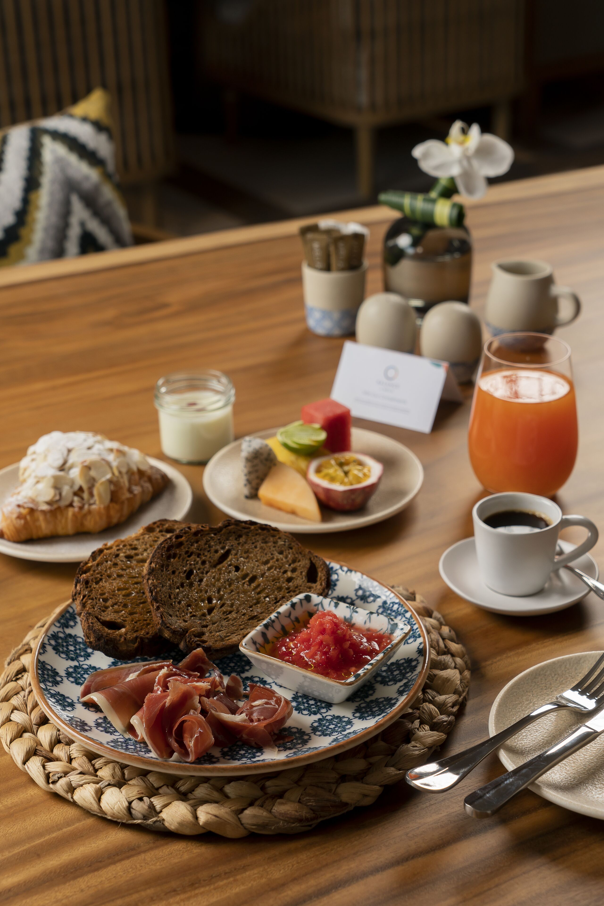 Ein Frühstückstisch mit Croissants, Brot, Käse, Schinken und einer Obstschale. Dazu stehen Kaffeetassen und Saft bereit, im Hintergrund steht eine Vase mit Blumen.
