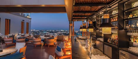 A rooftop bar with wicker furniture, a bar counter with bottles, and a city view.