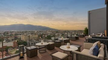 Una terraza en la azotea con muebles de mimbre, mesas redondas y vistas a un paisaje urbano y montañas.
