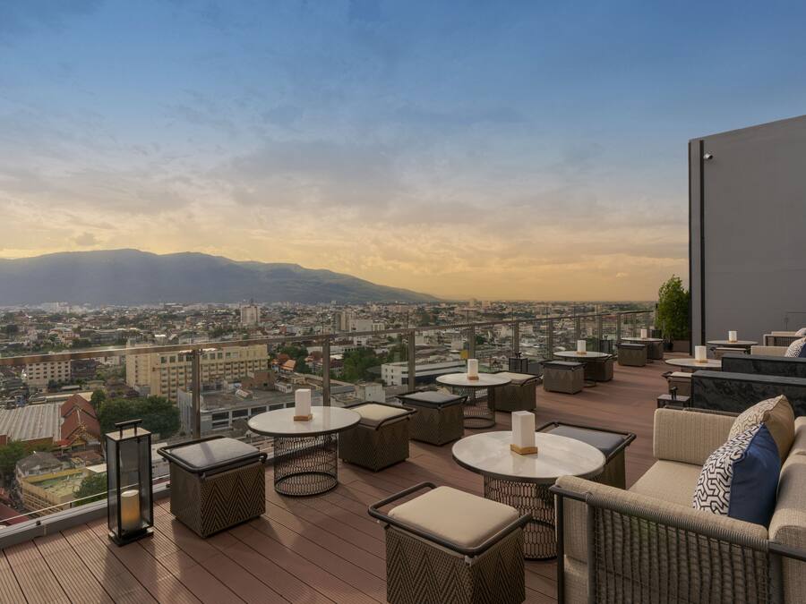 Una terraza en la azotea con muebles de mimbre, mesas redondas y vistas a un paisaje urbano y montañas.