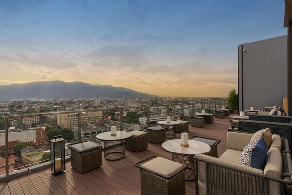 Una terraza en la azotea con muebles de mimbre, mesas redondas y vistas a un paisaje urbano y montañas.
