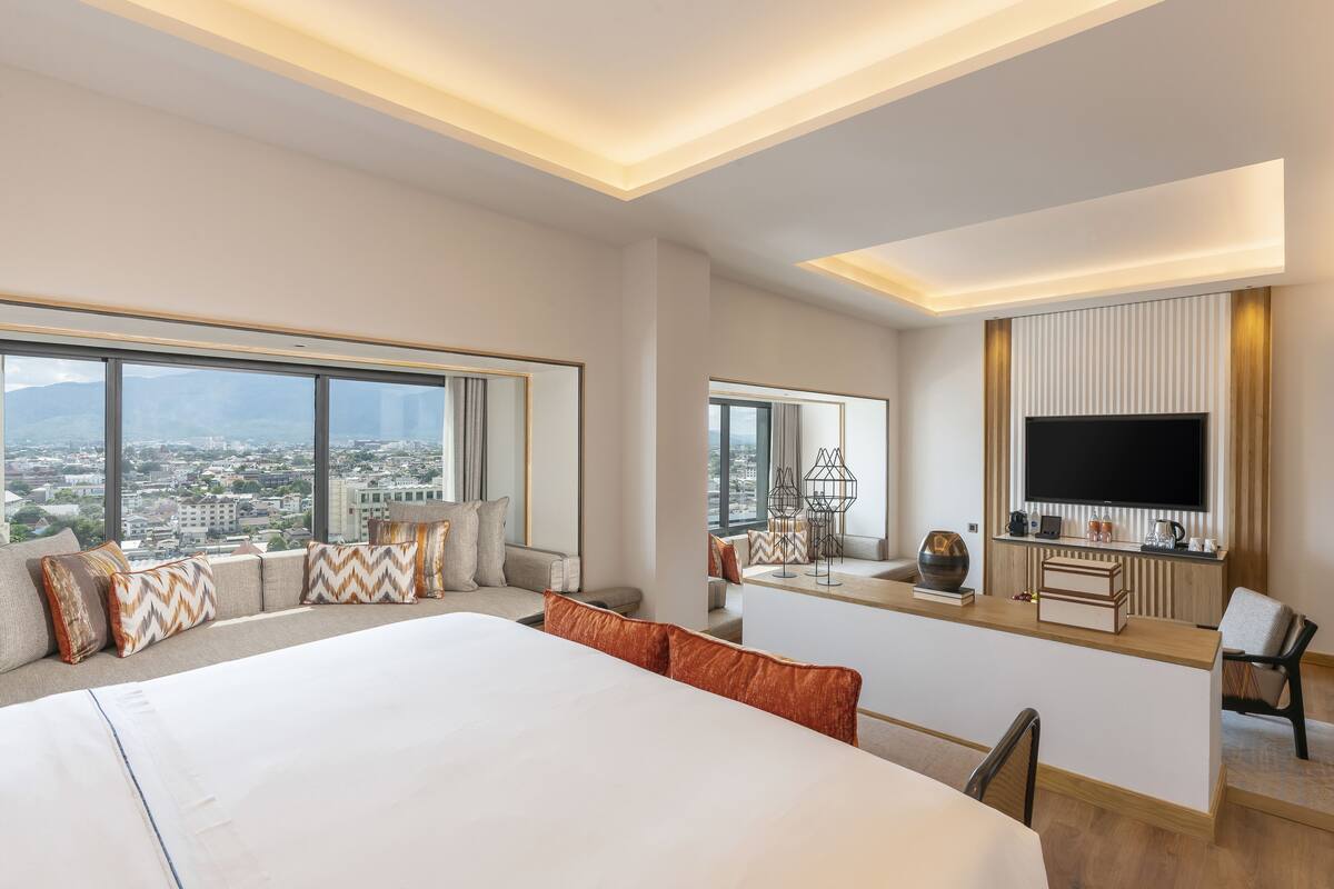 Ein modernes Hotelzimmer mit einem großen Bett, einem Schreibtisch mit Flachbildfernseher und Blick auf die Stadt durch große Fenster.