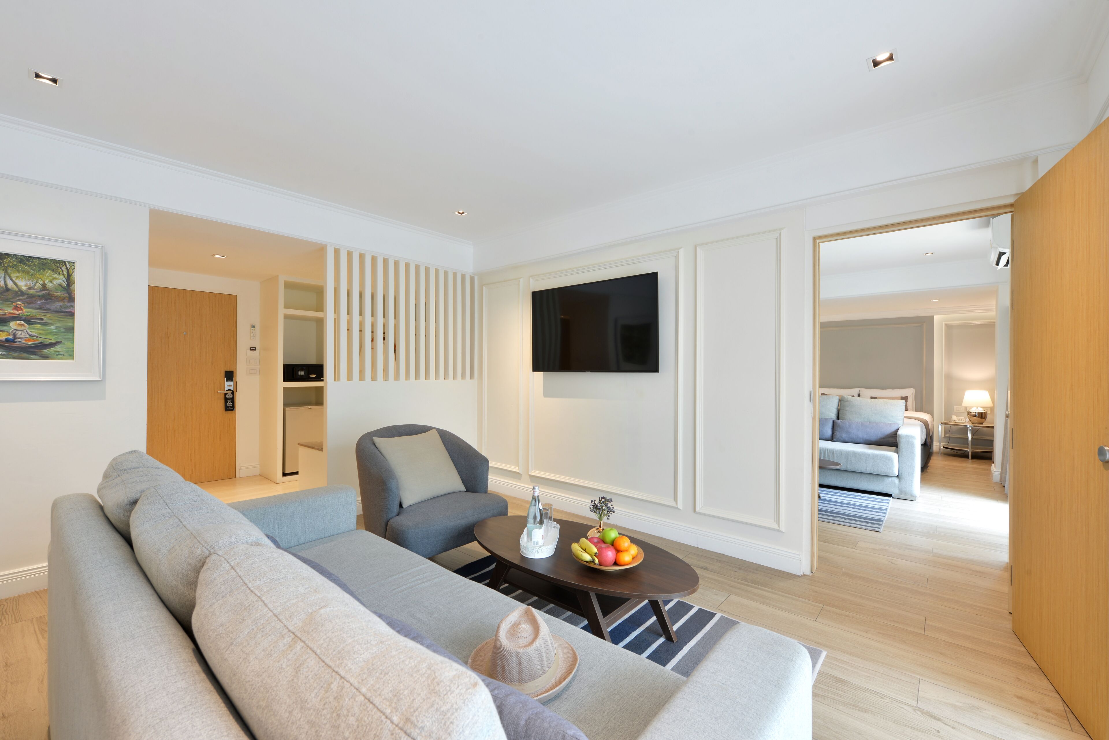 A modern living room with a grey sofa, a coffee table with fruit, a TV mounted on the wall, and a wooden door leading to another room.