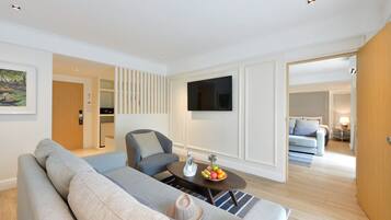 A modern living room with a grey sofa, a coffee table with fruit, a TV mounted on the wall, and a wooden door leading to another room.