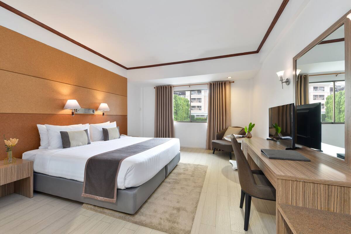 A modern hotel room with a large bed, a desk with a computer, a TV, and a view of buildings outside.