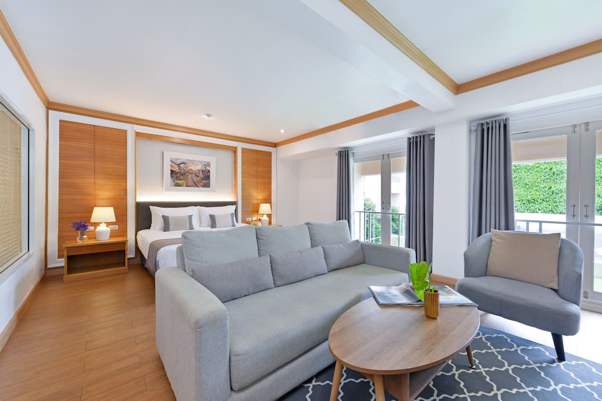 A modern hotel room with a sofa, a round coffee table, a bed with a headboard, a nightstand with a lamp, and a window with curtains.