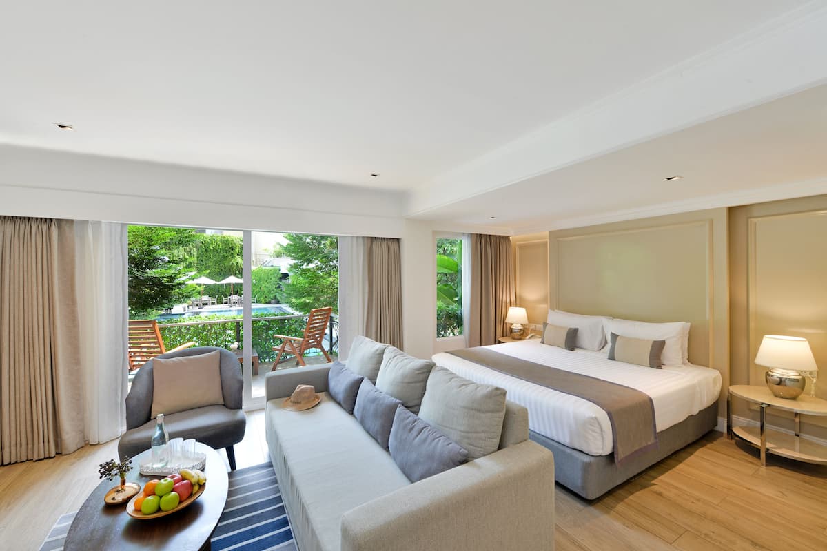 A hotel room with a large bed, a sofa, a dining table with fruit, and a view of the outdoors.