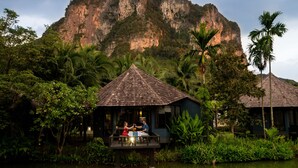 A bungalow with a thatched roof, surrounded by lush greenery and a body of water, with a large cliff in the background. - Peace Laguna Resort & Spa (Krabi)