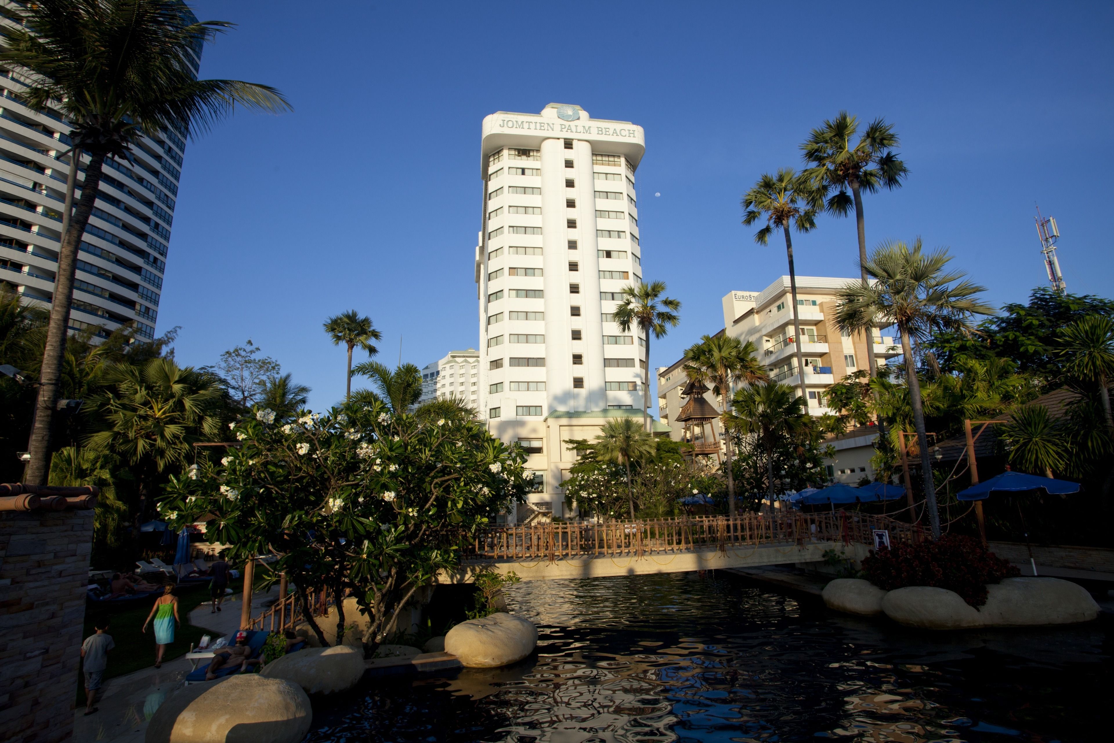Un hôtel doté d’une tour imposante sur laquelle on peut lire l’inscription « JOMTIEN PALM BEACH », ainsi que d’un élément décoratif aquatique composé de rochers et de palmiers.