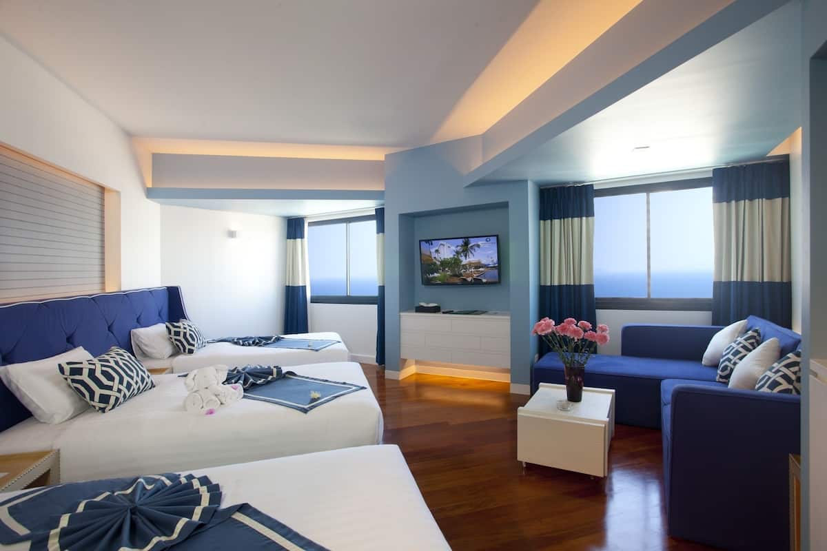 A hotel room with two beds, a sofa, a TV, and a view of the ocean.