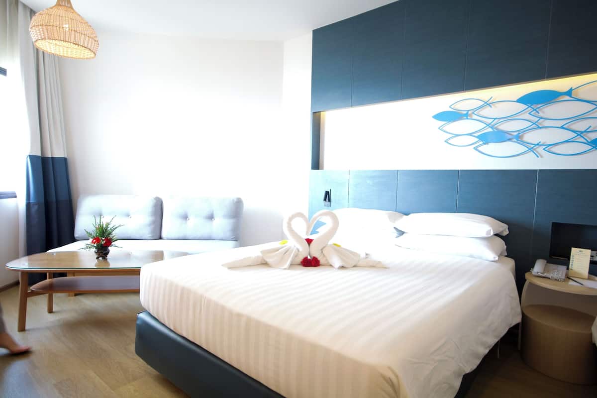 A hotel room with a large bed, a bedside table, a small table with a vase, a sofa, and a wall art piece.