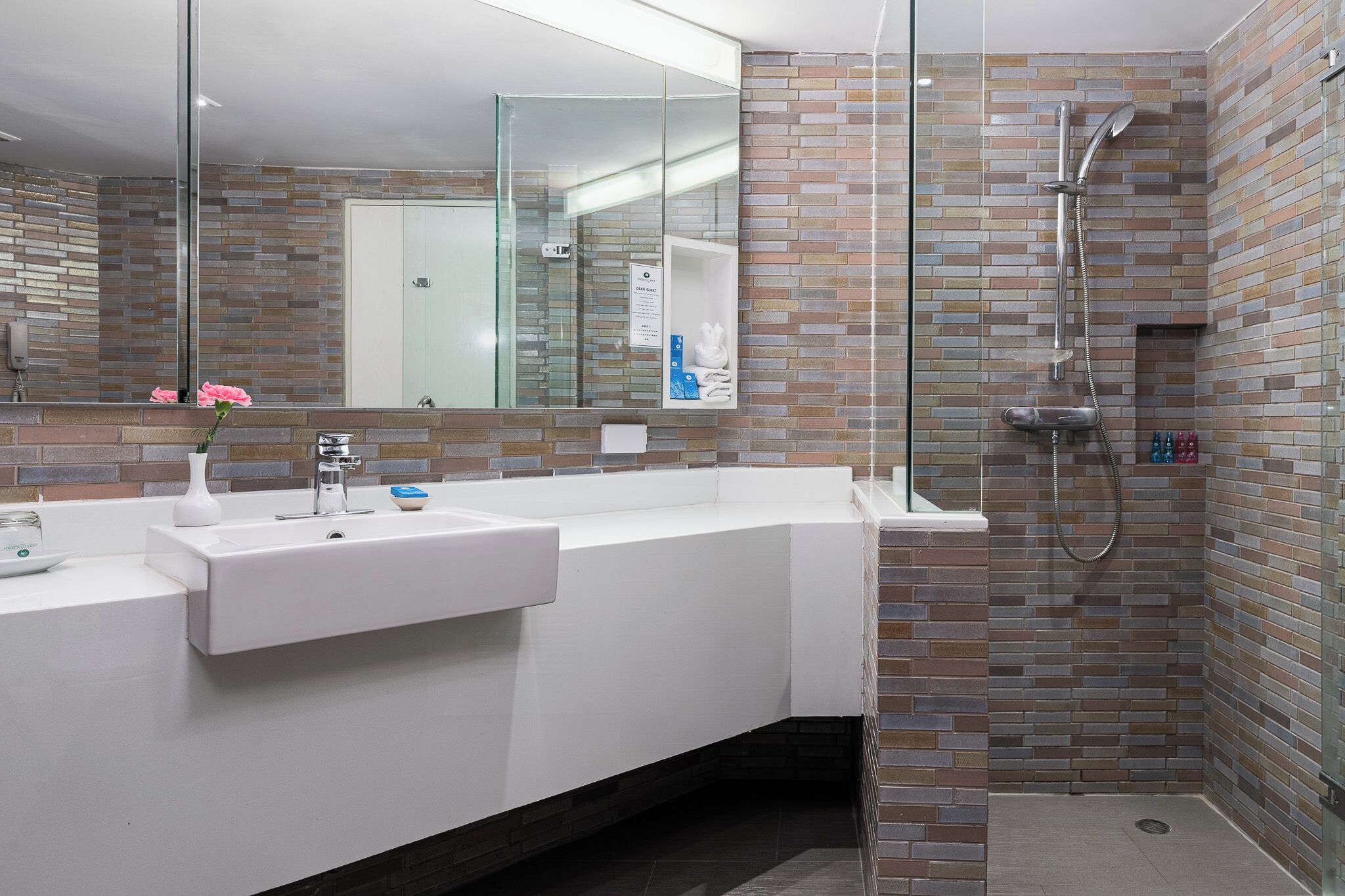 A modern bathroom with a white sink, a glass shower enclosure, and a mirror with a brick-patterned wall.
