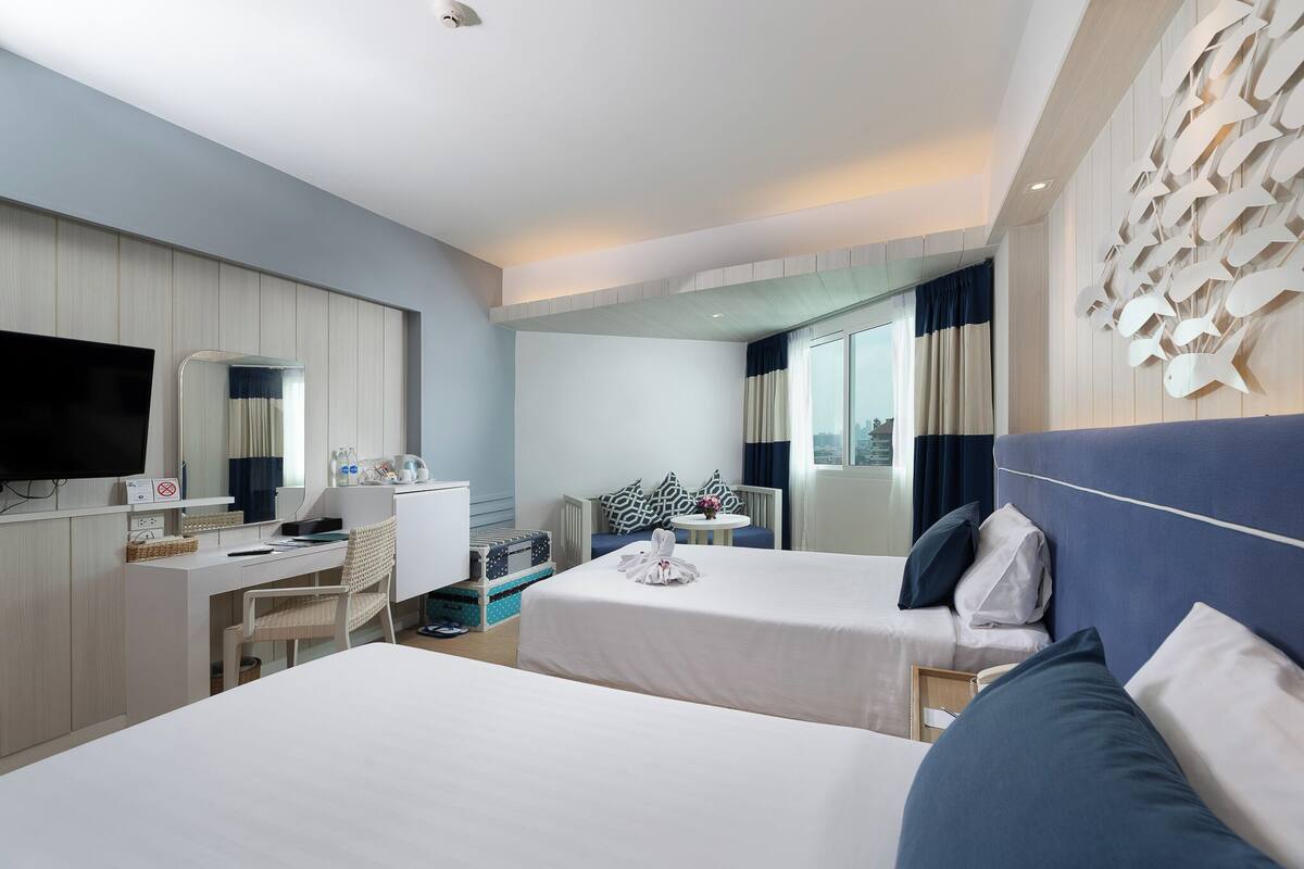A hotel room with two beds, a desk, a chair, a TV, and a large window with curtains.
