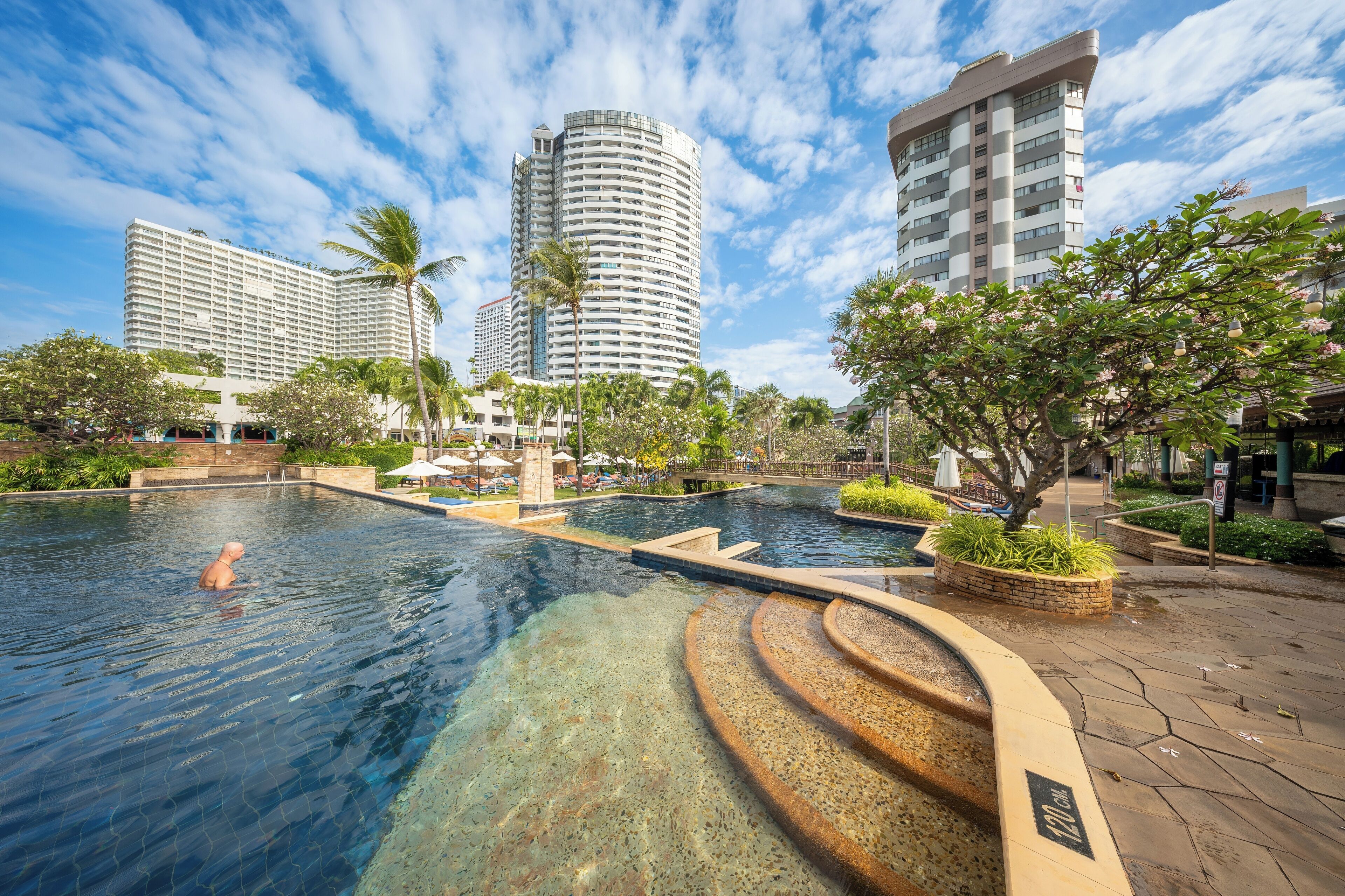 A swimming pool with a person swimming, surrounded by palm trees and modern buildings.