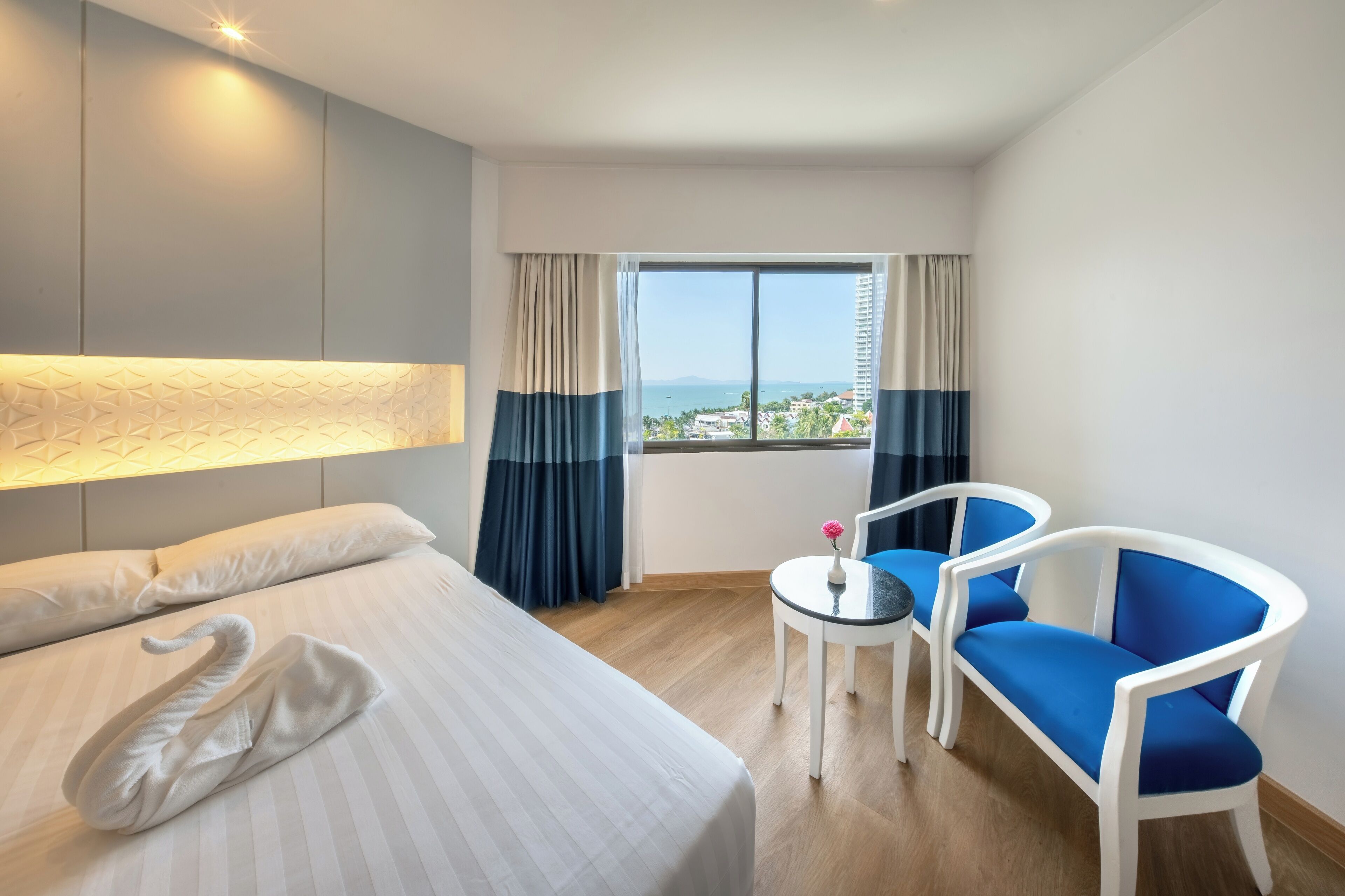 A hotel room with a bed, a small table with a vase, two blue chairs, and a window with curtains.