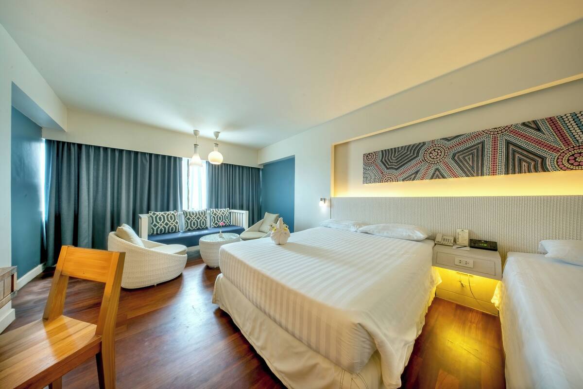 A modern hotel room with a large bed, a seating area with a sofa and chairs, a wooden chair, and a wall-mounted artwork.