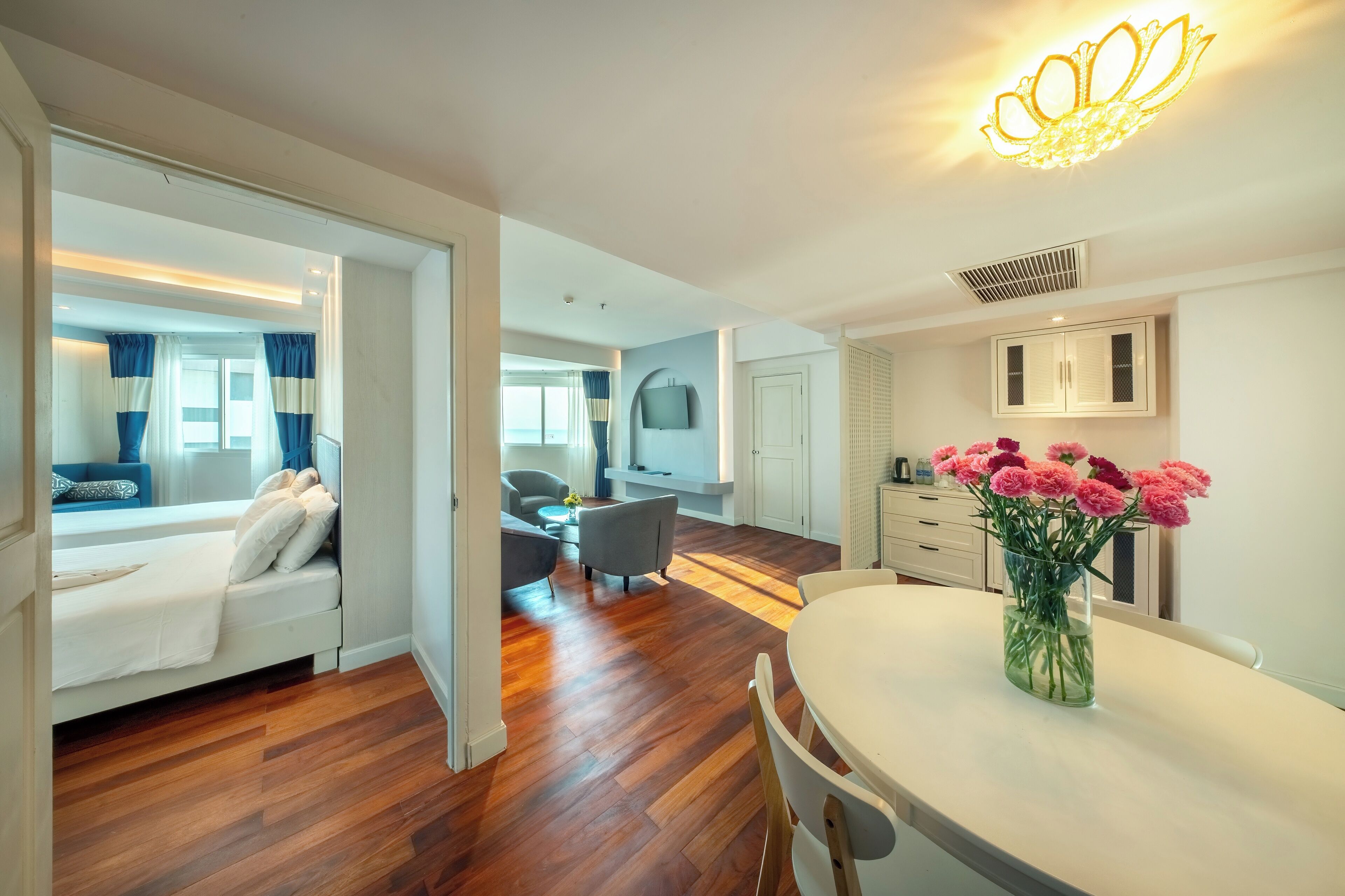 A modern hotel room with a bed, a dining area with a table and chairs, and a living area with a sofa and a TV.
