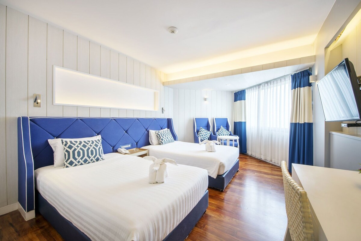 A hotel room with two beds, a TV, a desk, and a window with curtains.