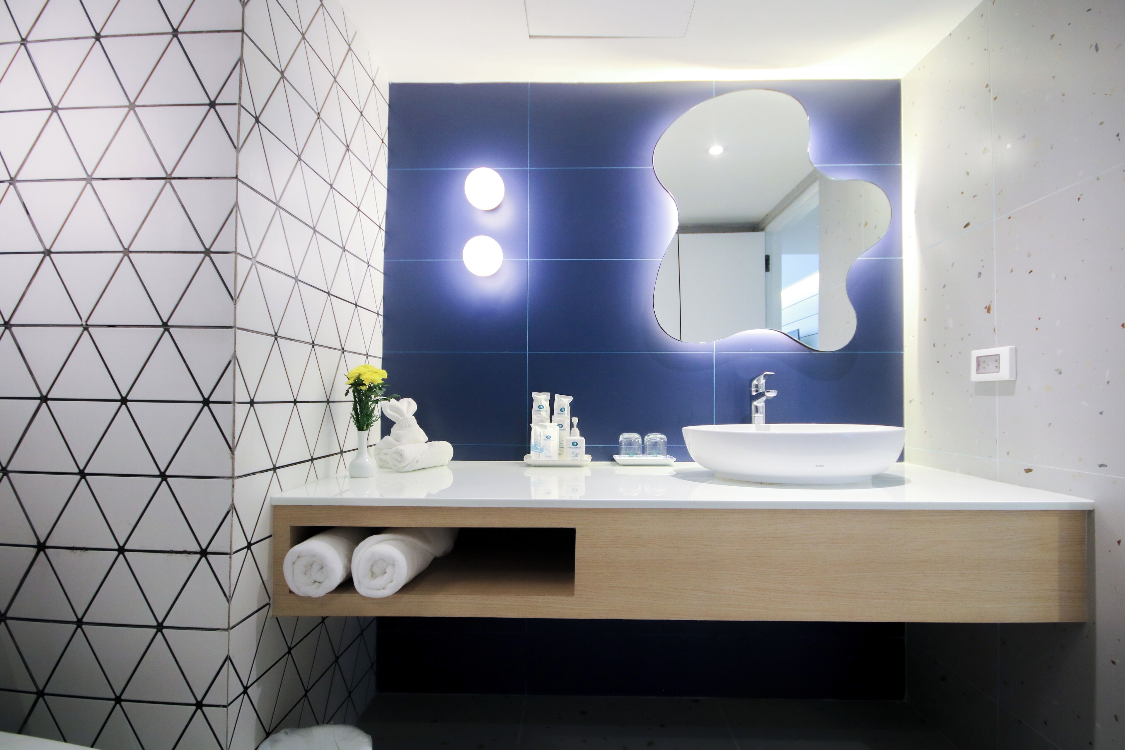 Uma casa de banho moderna com um painel de azulejos geométricos, um móvel de vaidade em madeira, uma pia branca e um espelho grande.