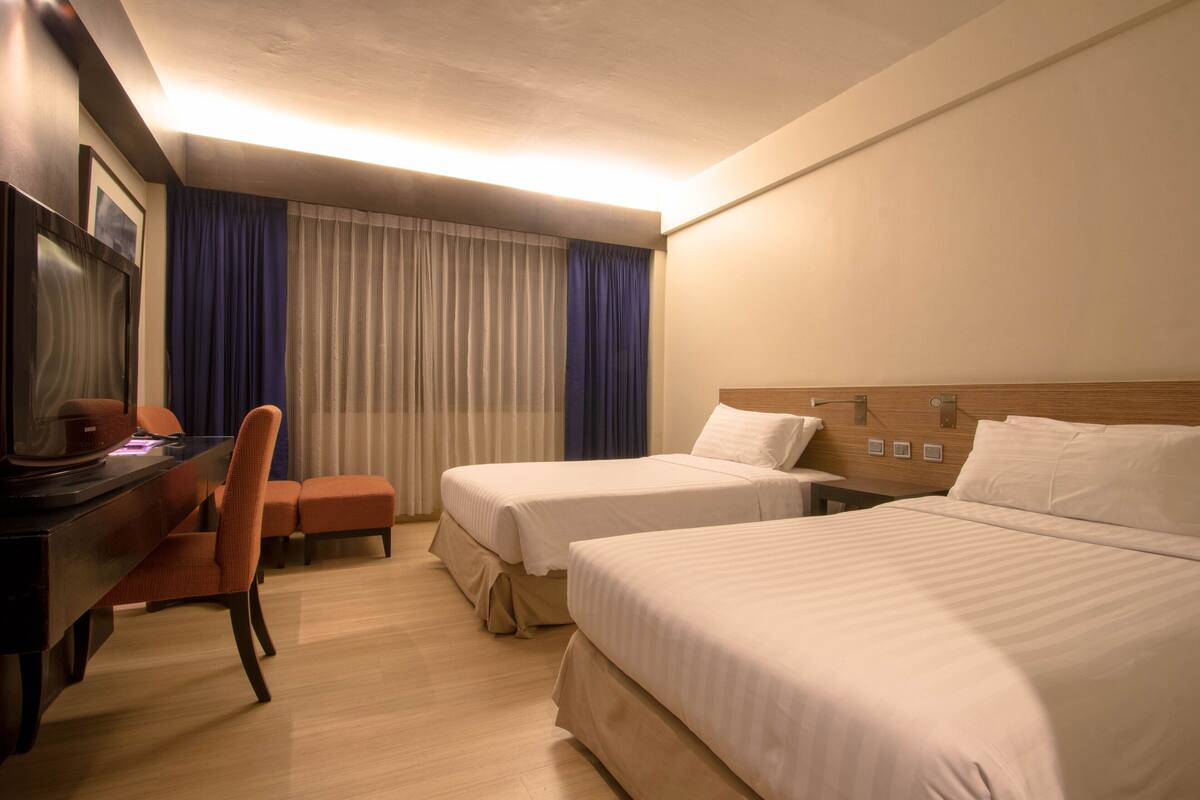 Une chambre d’hôtel avec deux lits simples, un bureau avec une chaise, une télévision et une fenêtre avec des rideaux.