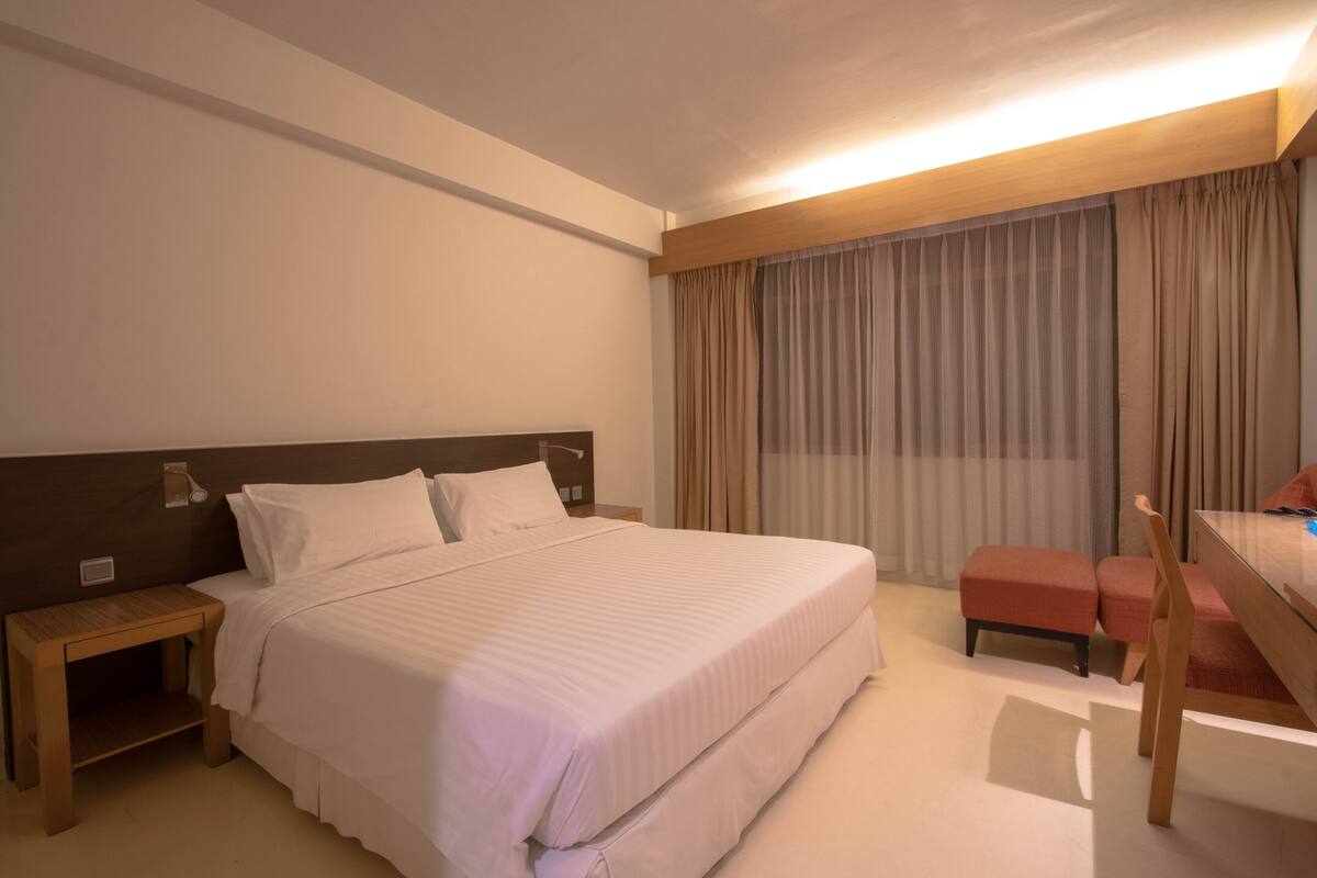 Une chambre d’hôtel avec un grand lit, une table de chevet en bois, un bureau avec une chaise et une fenêtre avec des rideaux.