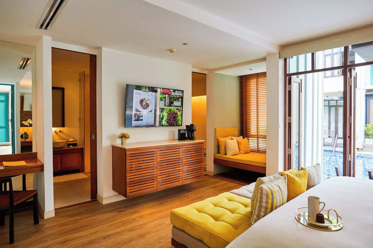 A modern hotel room with a wooden floor, a large bed, a sofa, a dining table, and a kitchen area.
