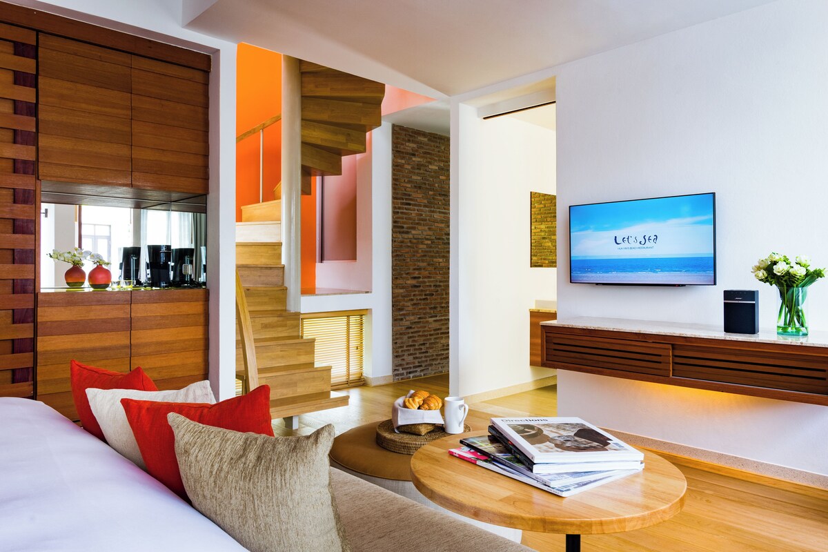 A modern living room with a wooden coffee table, a sofa with red and beige cushions, a television displaying 'Let's See', and a brick wall.