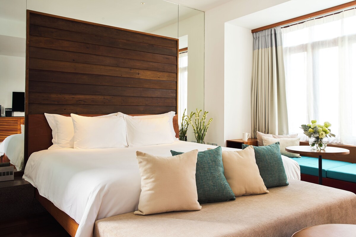 A modern hotel room with a large bed, a wooden headboard, a seating area with a table, and a view of the outdoors through large windows.