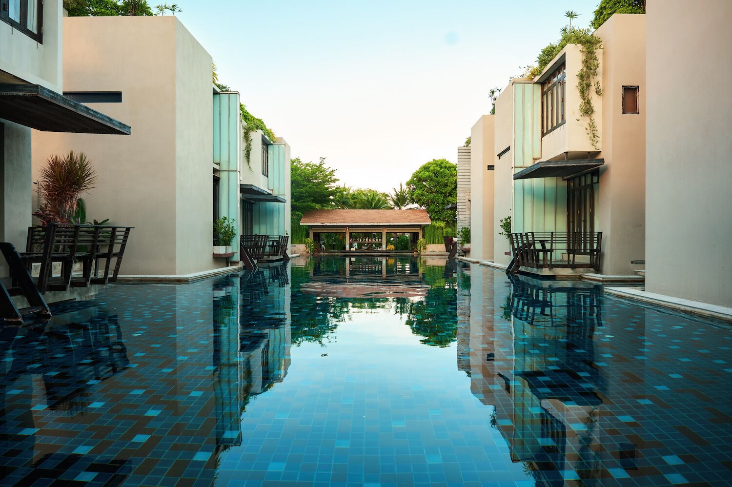 A swimming pool with a tiled floor, surrounded by buildings with a modern design, and a covered seating area in the center.