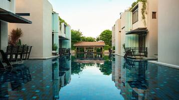 A swimming pool with a tiled floor, surrounded by buildings with a modern design, and a covered seating area in the center.