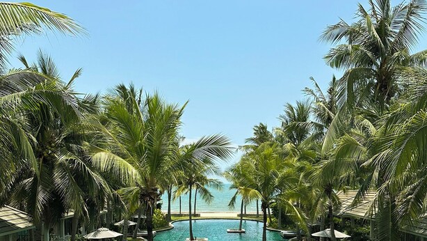 Ein Pool mit klarem Wasser, umgeben von Liegestühlen und Palmen, der zu einem Strand führt.