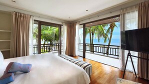 Ein Hotelzimmer mit einem großen Bett, einem Fernseher auf einem Schrank und einem Balkon mit Blick auf Palmen und das Meer.