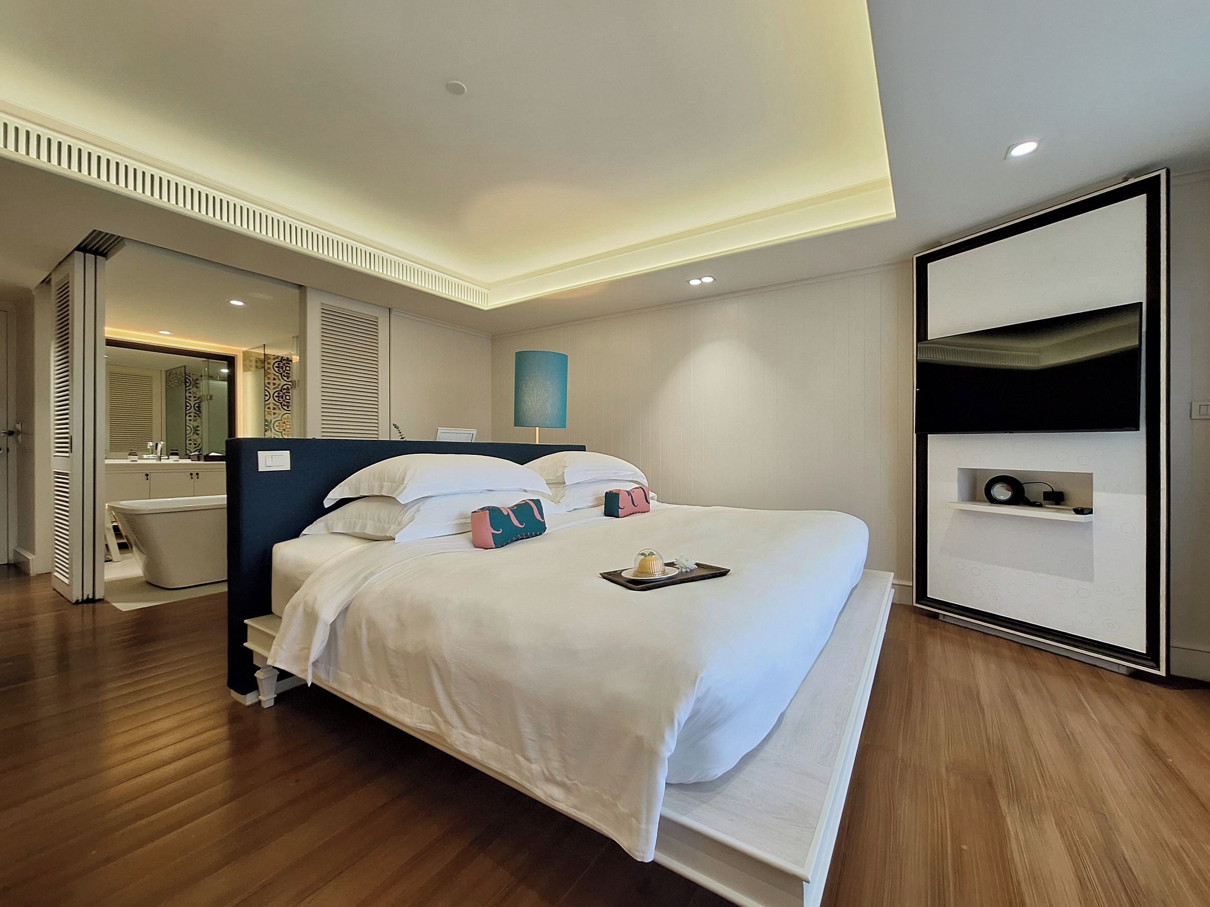 A modern hotel room with a large bed, a built-in desk, a vanity with a mirror, and a small shelf with toiletries.