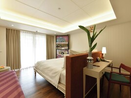 Habitación de hotel con cama, escritorio, silla, planta y un cuadro en la pared.