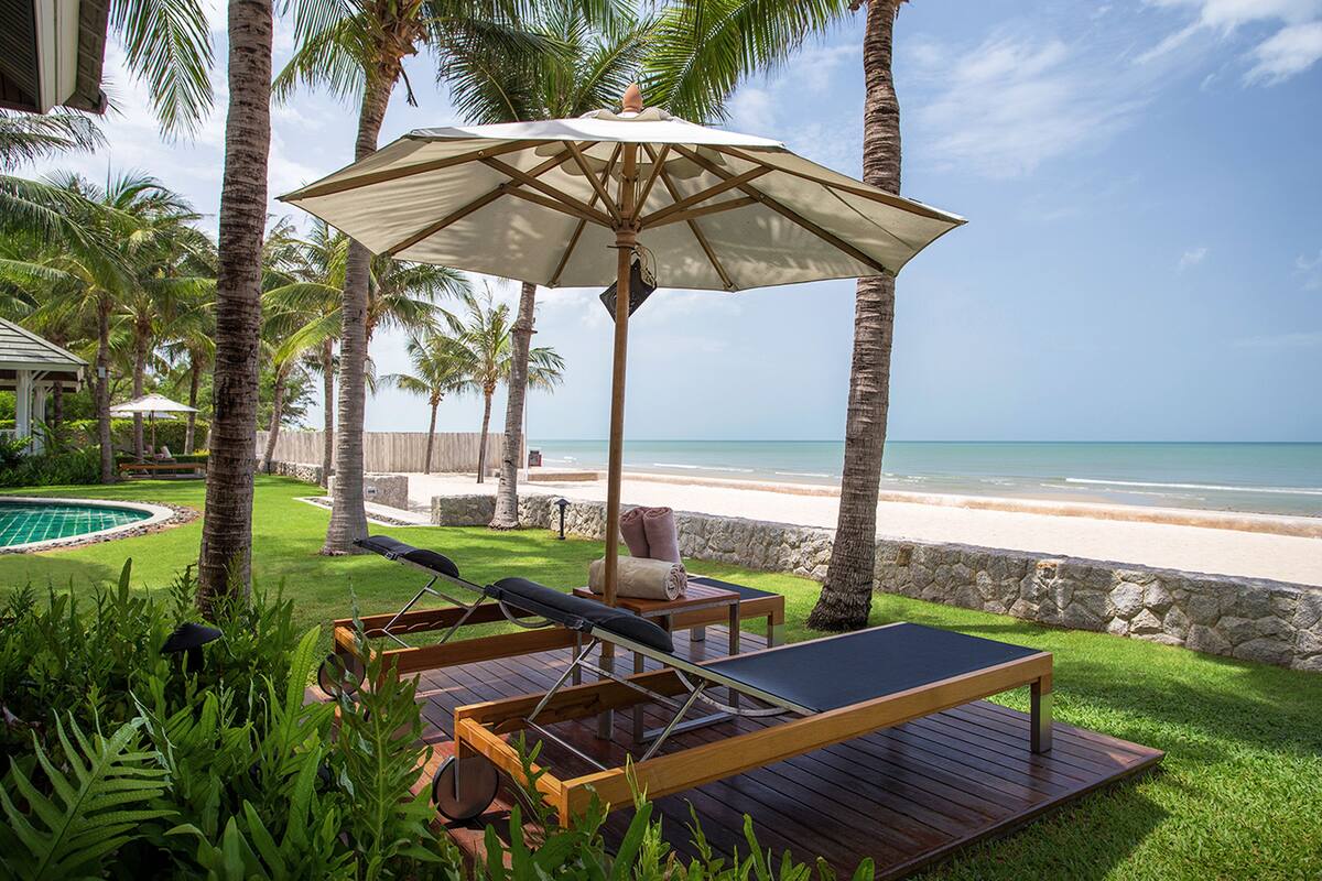Terraza de madera con una silla de descanso y una sombrilla, con vista a una playa con palmeras y una piscina.