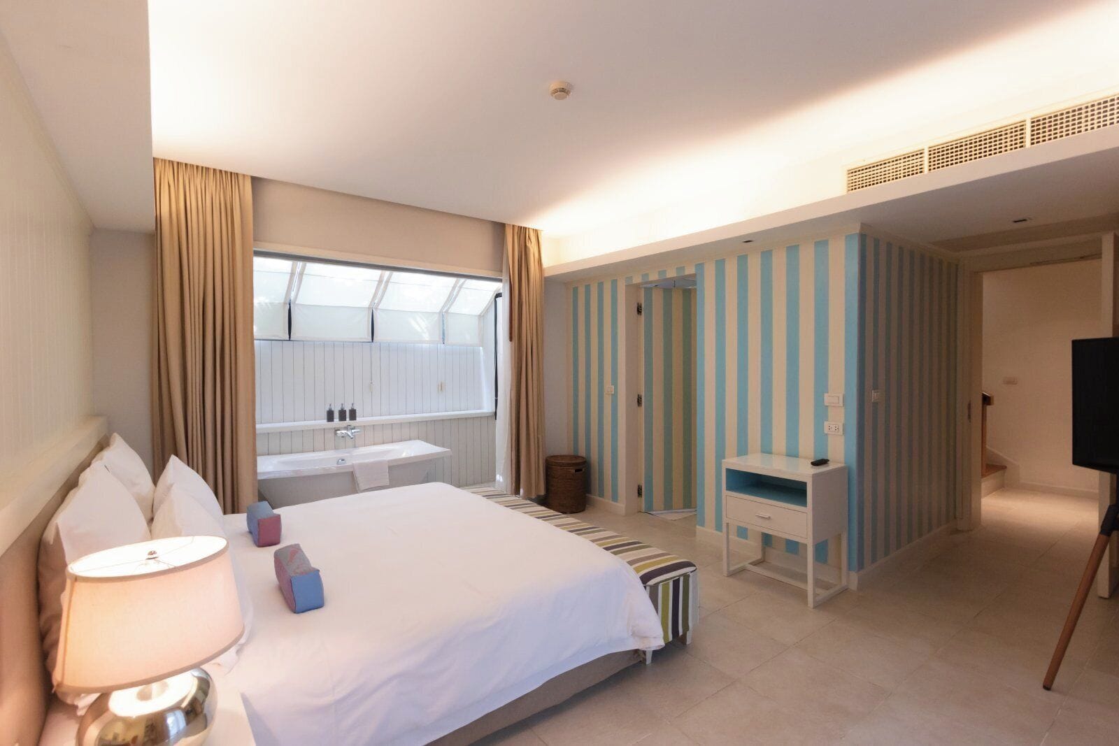 Habitación de hotel con cama, mesita de noche, lámpara, ventana con cortinas, baño con lavamanos y espejo, televisor sobre un mueble y paredes a rayas.