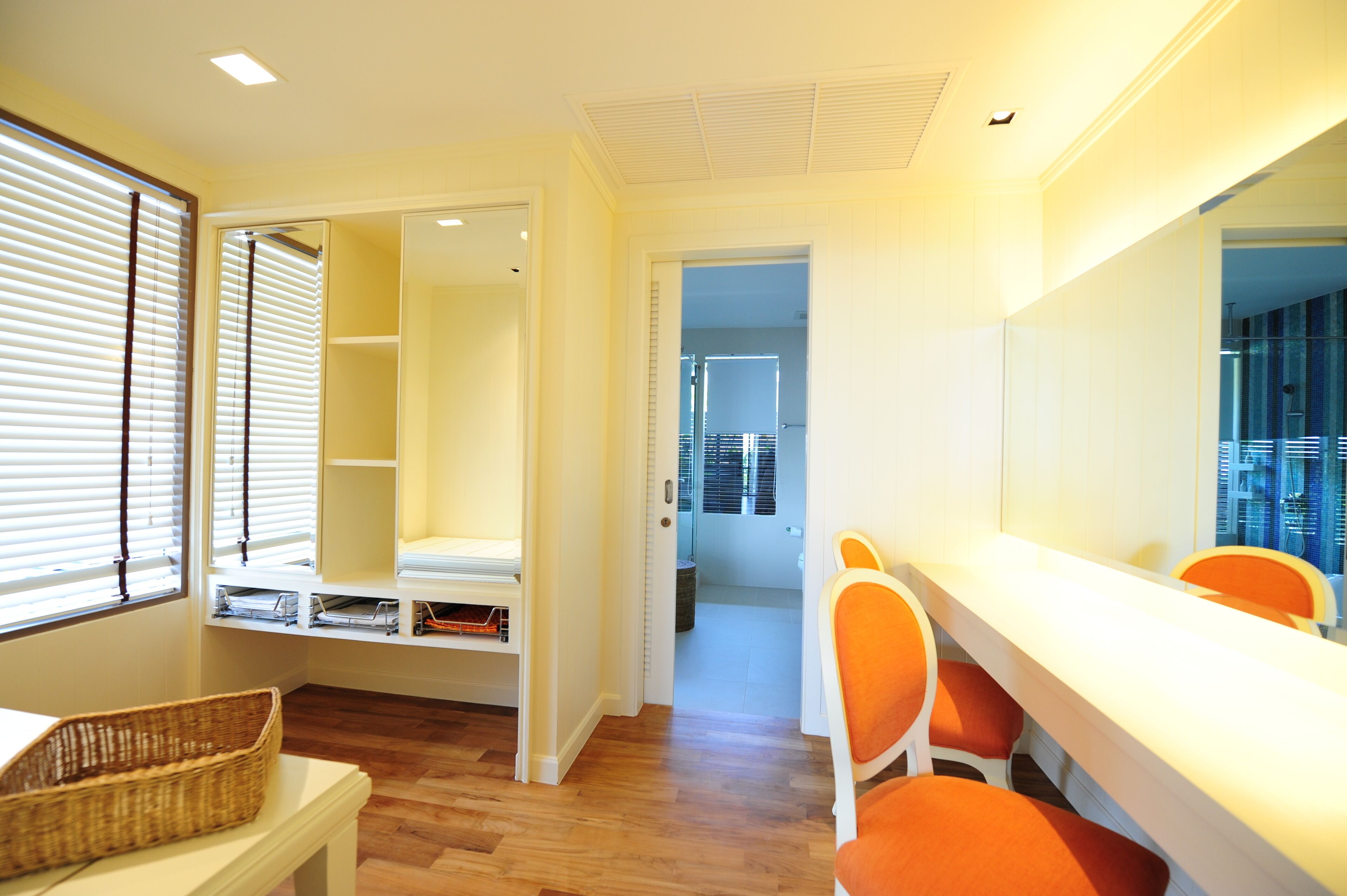 A room with a wooden floor, a white desk, orange chairs, a wicker basket, and a walk-in closet with shelves.