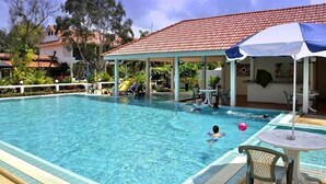 Una piscina con sillas de descanso y sombrillas, rodeada por una zona de estar cubierta y un edificio con techo de tejas rojas.