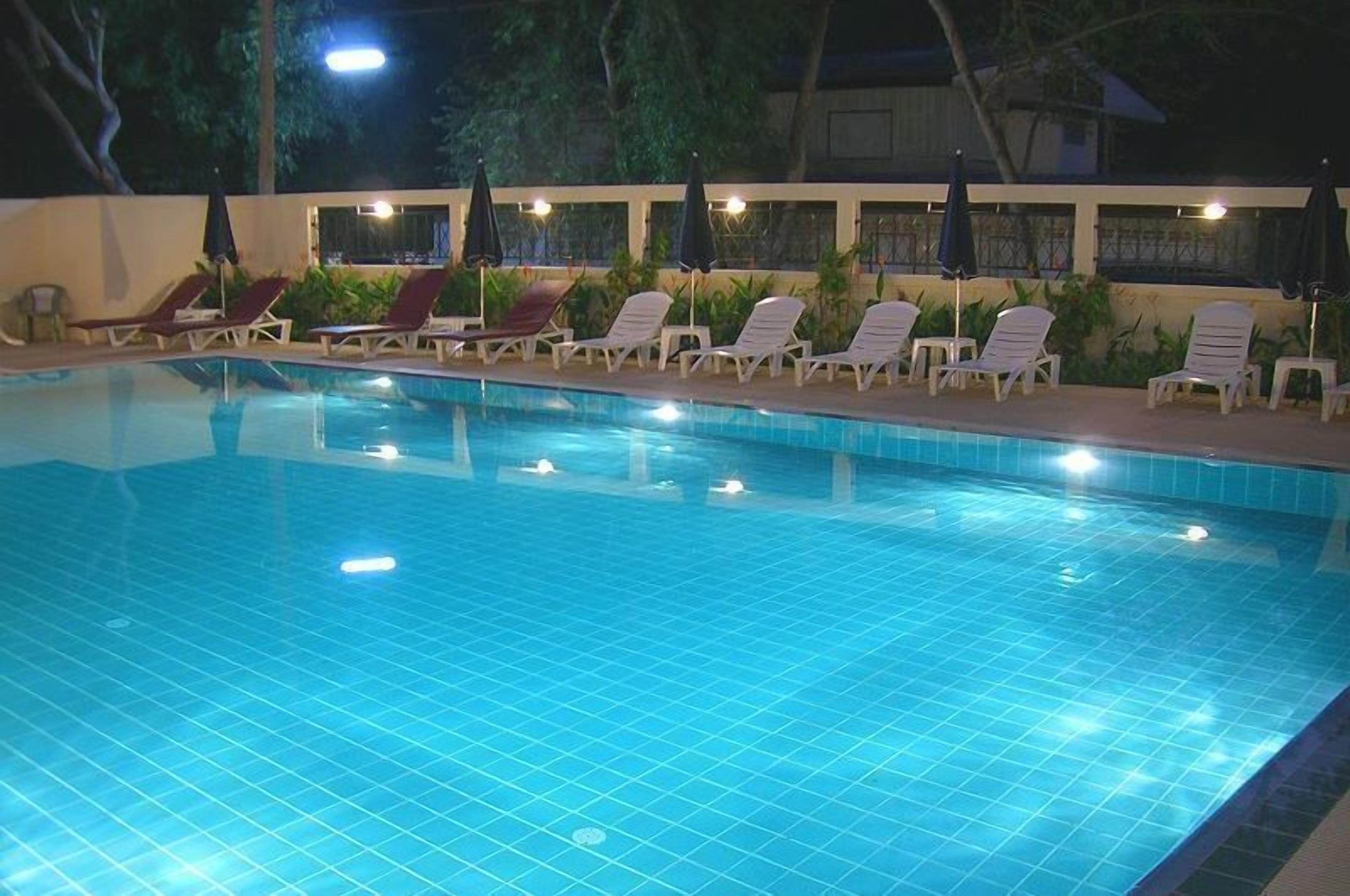 En swimmingpool med flisegulv, omgivet af hvide liggestole og parasoller, samt en væg med vinduer.