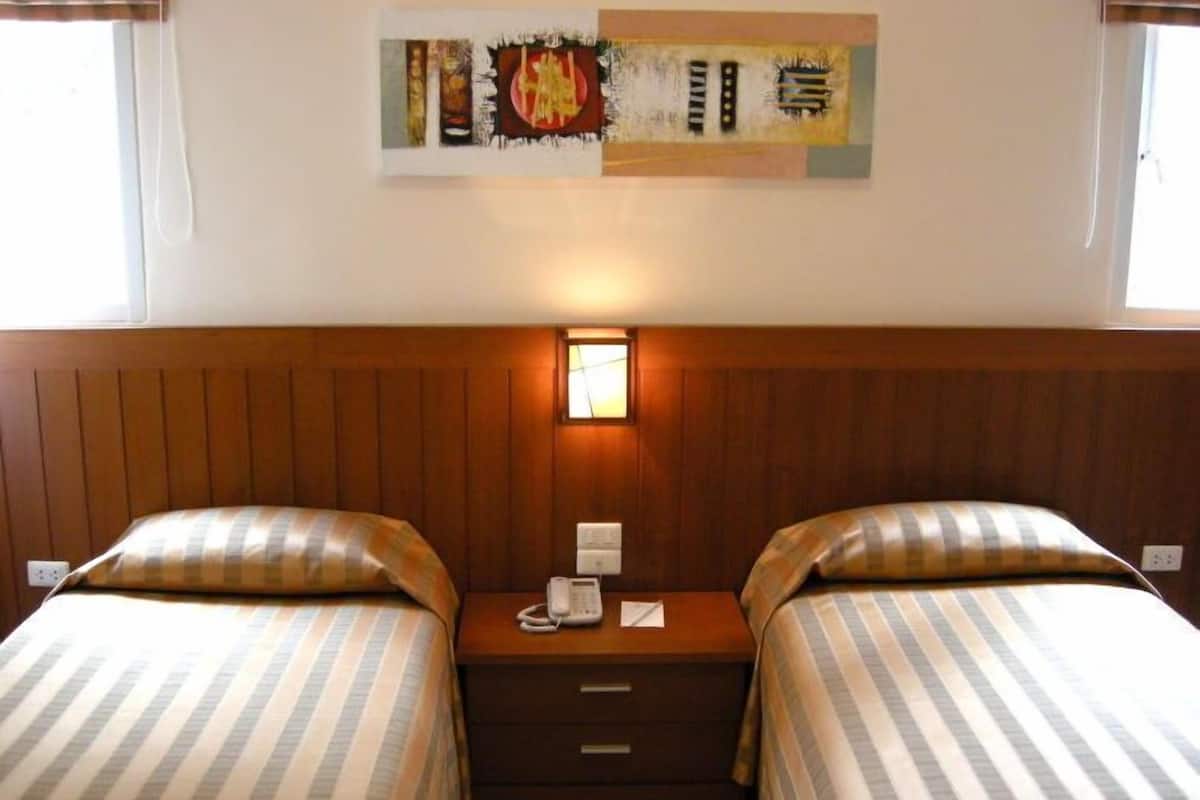 A hotel room with two beds, a wooden headboard, a bedside table with a phone, and a wall-mounted light.