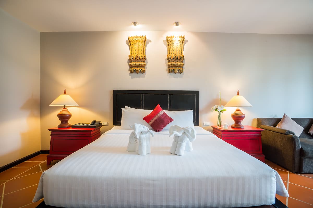 Ein Hotelzimmer mit einem großen Bett, zwei Nachttischen mit Lampen, einem roten Beistelltisch, einem roten Sessel und einem dekorativen Wandbild über dem Bett.