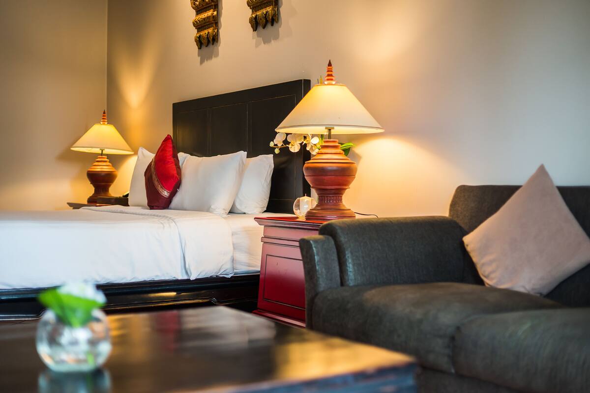 Ein Hotelzimmer mit einem Bett, zwei Nachttischlampen, einem roten Nachttisch, einem grauen Sofa und einem kleinen Tisch mit einer Vase.