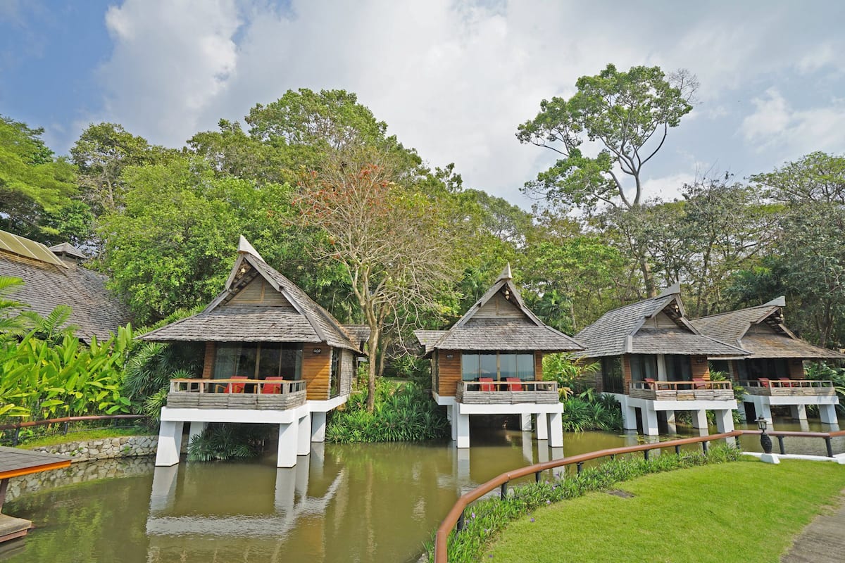 A row of wooden overwater bungalows with balconies, surrounded by lush greenery and a calm water body.